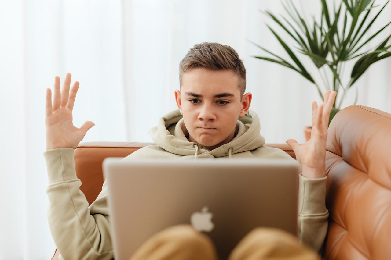 Teen sitting on couch with laptop, appearing frustrated, indoors.